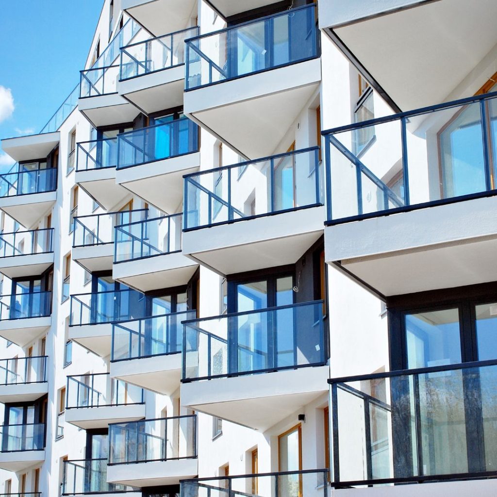 Modern white apartment building with multiple glass balconies and large windows under a bright blue sky.